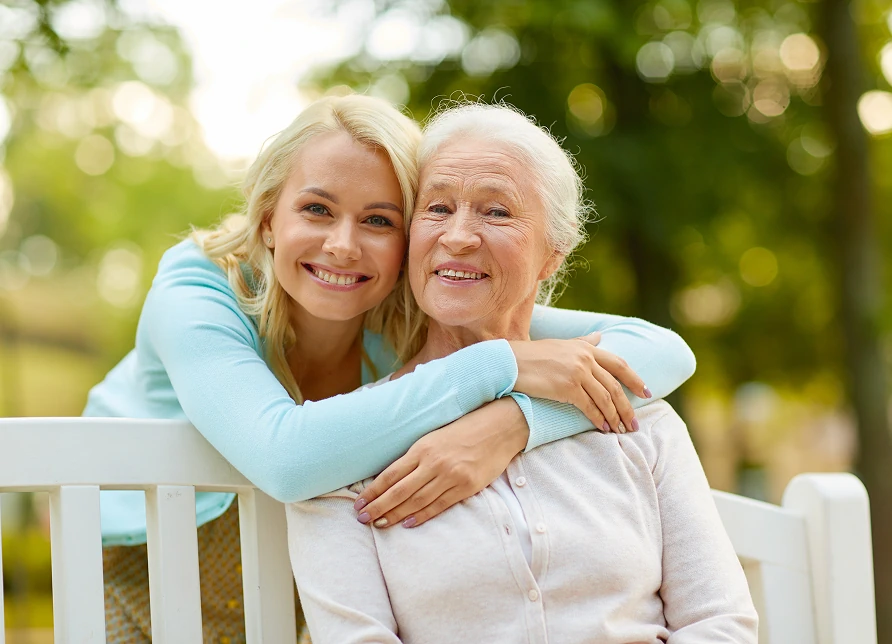 Blond kobieta przytulająca od tyłu starszą panią siedzącą na ławce w ogrodzie